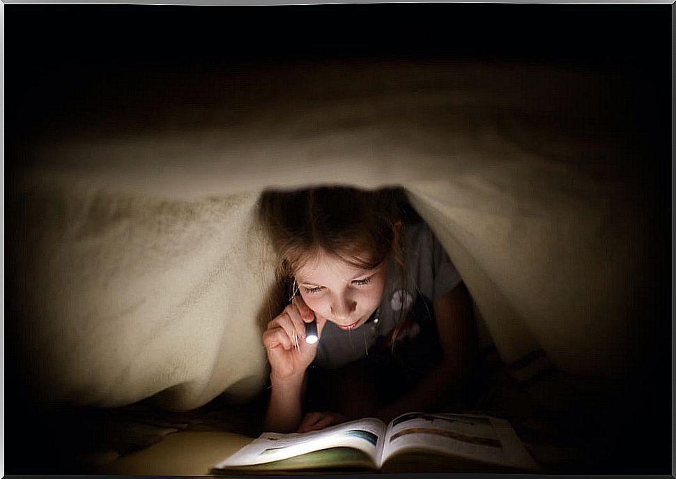 Girl reading one of the stories in the collection Look below!  under the covers of his bed with a flashlight.